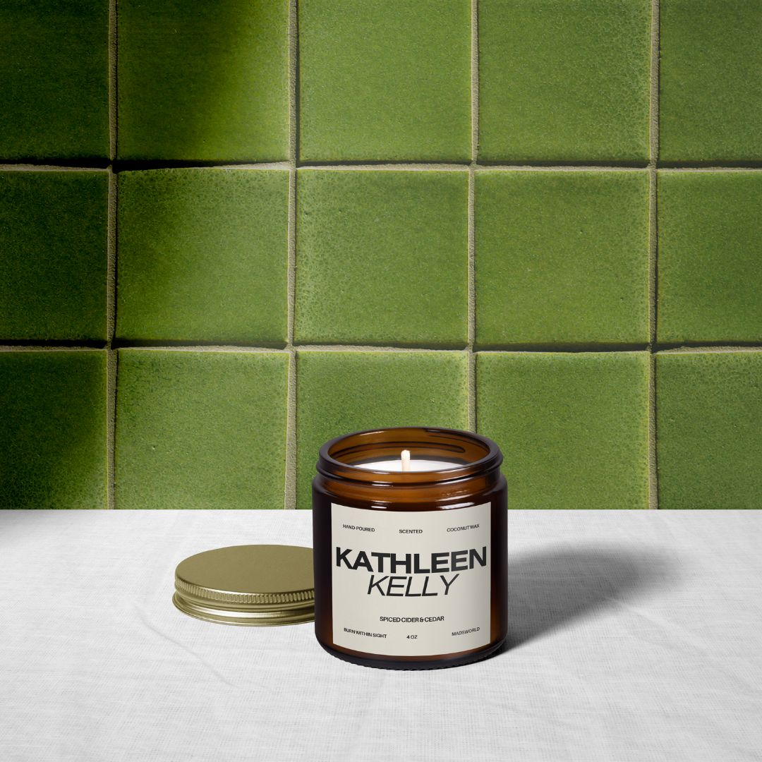 A brown glass candle jar labeled Kathleen Kelly sits on a white cloth with its gold lid off, against a backdrop of green tiled walls. The candle is lit.