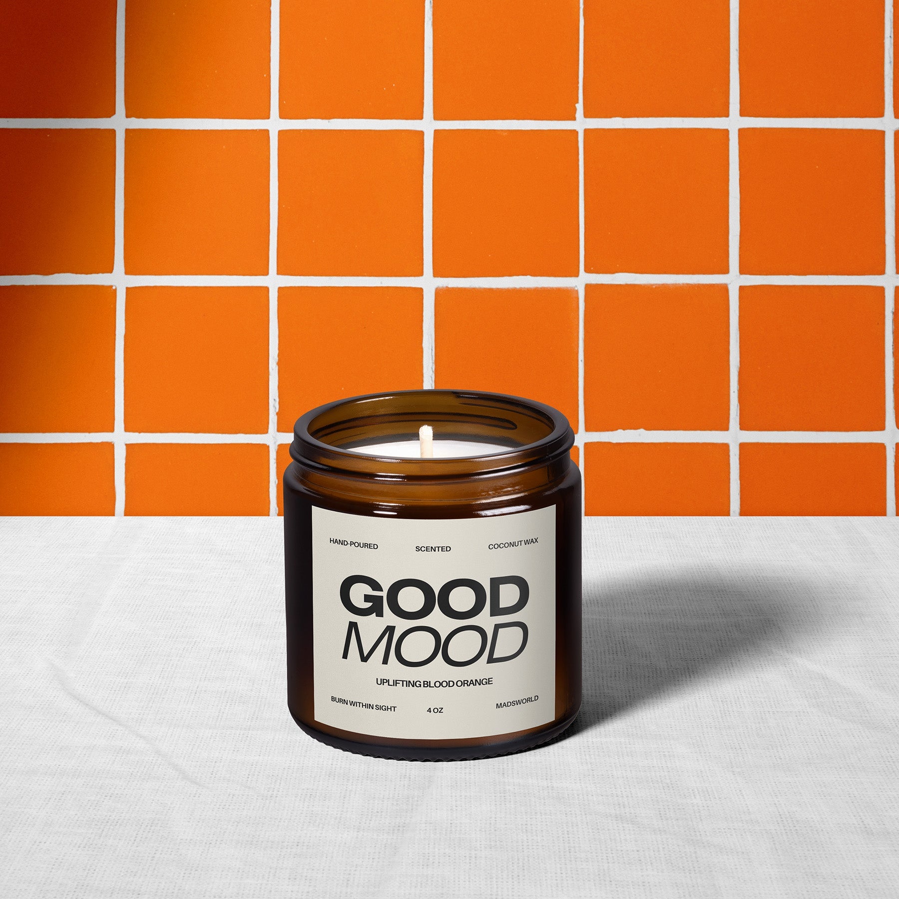A brown glass candle labeled GOOD MOOD sits on a white cloth, with a bright orange and white tiled wall in the background.